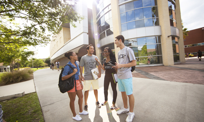 Four University of Central Florida students wearing UCF shirts talk and laugh outside a modern campus building.