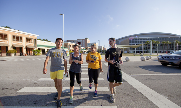 Four UCF students in branded T-shirts walk together across a crosswalk near campus buildings.